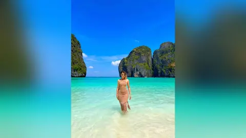 Woman in a beige halter dress standing in turquoise tropical ocean water by limestone cliffs under a blue sky.