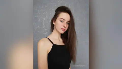 Young woman with long brown hair wearing a black tank top against a textured gray studio background.