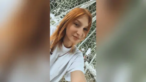 Redheaded woman in a white t-shirt smiling outdoors against a white geometric lattice background.