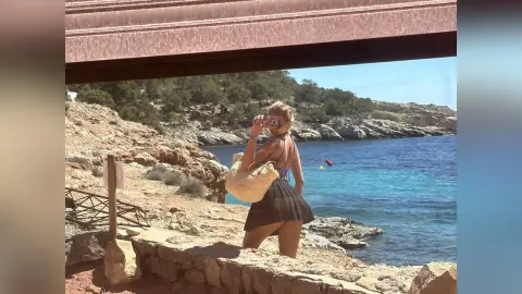 Woman in a black bikini and straw bag enjoying a sunny summer day by the rocky Mediterranean coastline.
