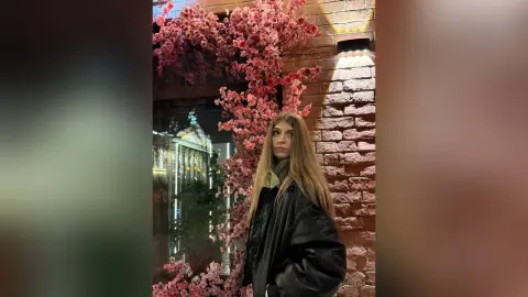 Young woman in a black leather jacket standing by a pink floral window display at night.