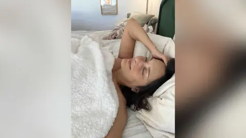 MichaelaBelle lies on a white bed, resting her head on her hand while covered in a white fuzzy blanket.