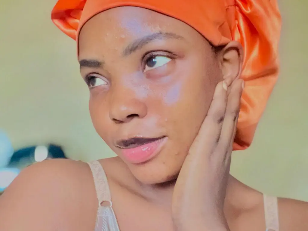 Young woman with glowing skin wearing an orange satin hair bonnet in a soft, natural indoor setting.
