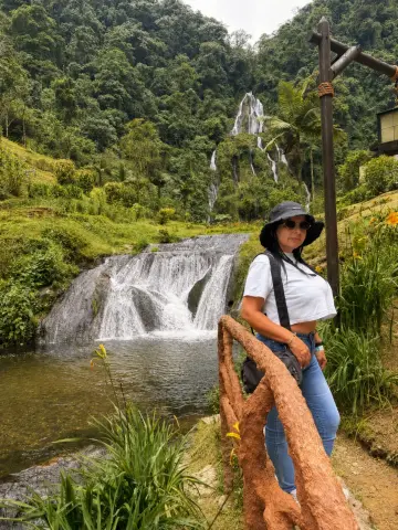 Victoria_Velvet stands by a wooden railing overlooking a lush waterfall and tropical forest scenery.