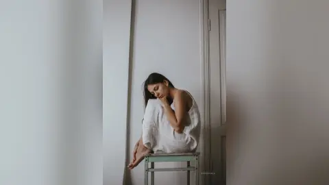Melancholic young woman in a white tank top sitting on a stool against a minimalist white wall.