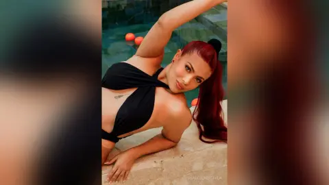 IsabellaEstrada poses in a black cut-out bodysuit with her red hair in a high ponytail, leaning against a stone ledge by a pool.