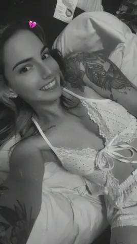 Smiling young woman with tattoos wearing white lace lingerie while relaxing on a bed in a moody black and white photo.