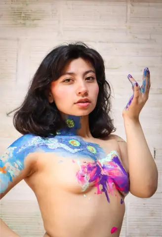 Young woman with colorful body paint posing against a white brick wall in a creative artistic mood.