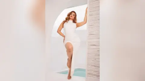 Elegant woman in a white sleeveless maxi dress posing in a bright, minimalist modern white interior.