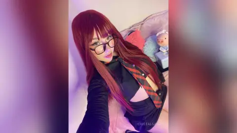 Young woman with long red hair and glasses wearing a black outfit in a cozy bedroom setting.