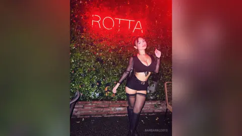 Glamorous woman in a black crop top and shorts posing under a red neon sign against a lush green wall.