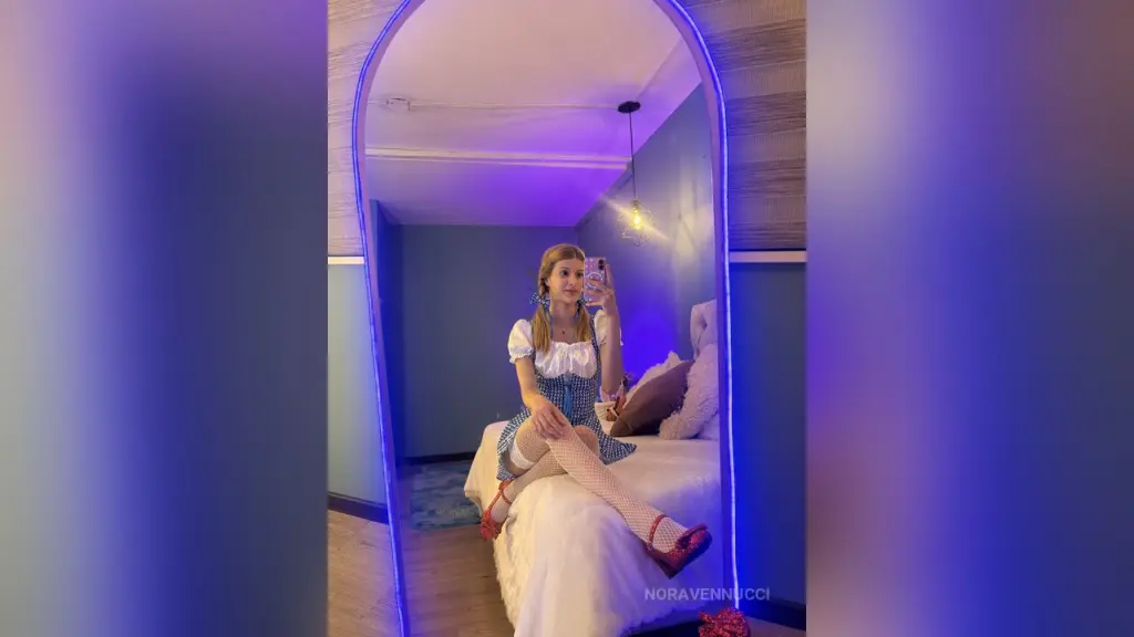 NoraVennucci — Young woman in a blue gingham dress sitting on a bed in a room with moody purple neon lighting.