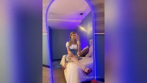 Young woman in a blue gingham dress sitting on a bed in a room with moody purple neon lighting.