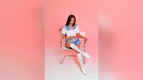 MilanaVolkova wearing a white and blue plaid schoolgirl outfit and long white socks, sitting on a white chair against a pink background.