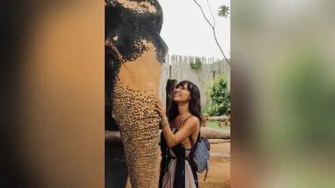 Smiling woman in a striped dress petting a large elephant at a sunny wildlife sanctuary.