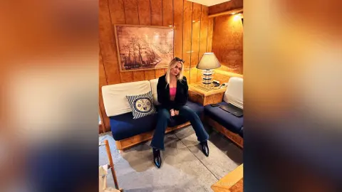 MilenaBia sits on a blue sofa wearing a black jacket and blue jeans in a wood-paneled room.