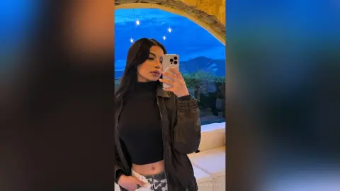 Young woman in a black turtleneck and leather jacket taking a selfie against a scenic mountain view at dusk.