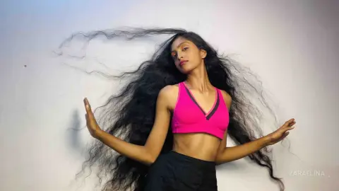 Young woman with long flowing hair wearing a pink sports bra and black shorts against a white background.