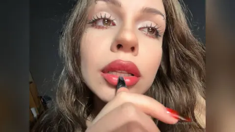 Young woman with long wavy hair and facial piercings applying red lipstick in a close-up portrait.