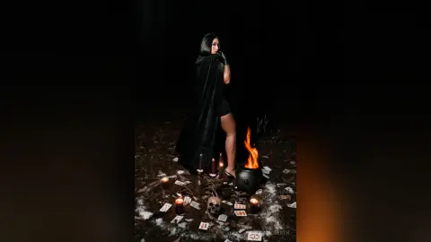 KimRoberth poses in a black cape and dark outfit, surrounded by lit candles, a skull, and tarot cards on a dark floor.