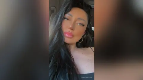 Glamorous woman with long dark hair and pink lipstick posing in a car for a chic selfie.