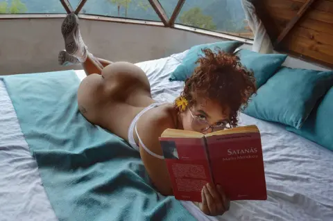 Naked woman with curly hair reading a red book on a bed in a bright, sunlit room.