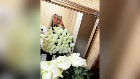 Blonde woman in black outfit holding a massive bouquet of white roses in a mirror selfie.