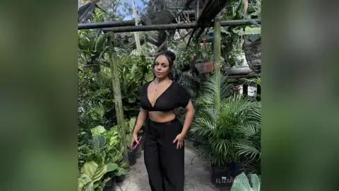 Stylish woman in a black crop top and trousers posing in a lush, sunlit tropical plant nursery.