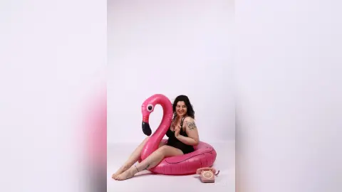 KiraKovall wearing a black bodysuit, sitting on a large pink inflatable flamingo against a white background.