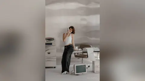 Young woman in a white tank top and black jeans sitting in a bright, minimalist modern office setting.
