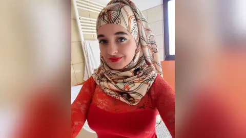 Smiling woman in a red lace top and patterned beige hijab posing in a bright indoor setting.