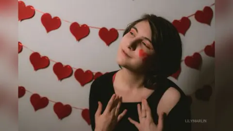 Young woman in a black top smiling against a backdrop of red heart decorations for a romantic Valentine's mood.