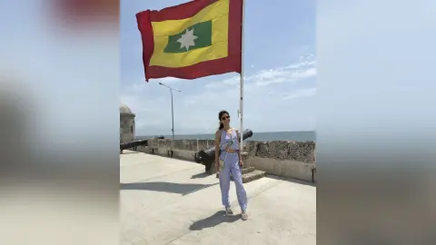 Stylish woman in light blue outfit standing by a historic stone fort under a bright sunny sky with a large flag.