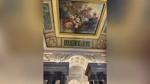 Classical ceiling mural painting in a grand museum hall with ornate gold architectural details.