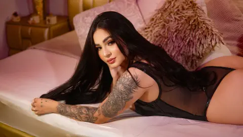Young woman with long dark hair in a black bodysuit relaxing on a bed in a cozy, warm-lit bedroom.