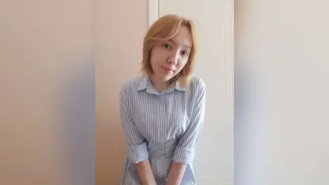 Young woman in a blue striped button-down shirt posing in a bright, minimalist indoor setting.