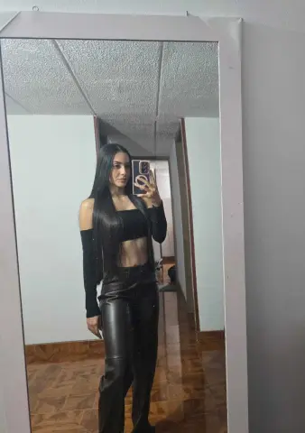Young woman in black crop top and leather pants taking a mirror selfie in a modern room.