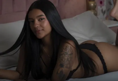 Young woman with long dark hair in black lingerie relaxing on a white bed in a moody, intimate setting.