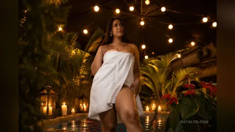 NatalyBloom poses in a white towel by a pool at night under warm string lights.