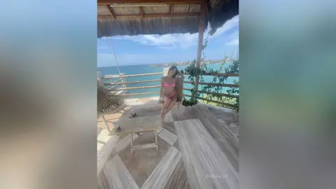 Young woman in a pink bikini relaxing on a wooden tropical deck overlooking a turquoise ocean under a sunny sky.
