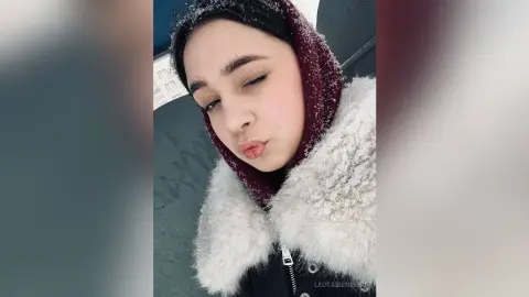 Young woman in a cozy white faux fur coat and burgundy hood smiling in a snowy winter setting.