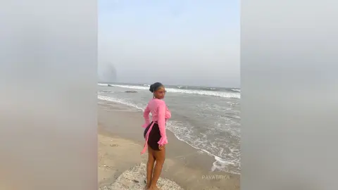 Young woman in a pink long-sleeve top standing on a sandy beach under a cloudy sky.