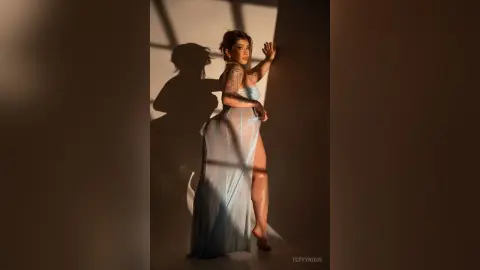 Tattooed woman in a flowing light blue sheer gown posing in dramatic sunlight against a shadowed wall.