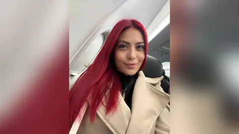 Smiling woman with long red hair wearing a beige coat sitting on an airplane.