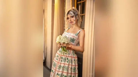 Blonde woman in a floral white summer dress holding a bouquet of white flowers by a classic building pillar.