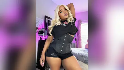 BossyBella — BossyBella poses in a black corset and black shorts, showing off her blonde hair and curves in a bedroom setting.