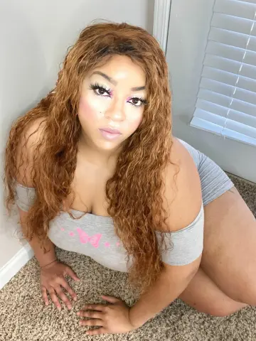 Curvy woman with long wavy ginger hair wearing a grey off-the-shoulder top sitting on a carpeted floor.