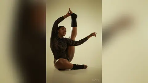 KeianHoz poses in a black mesh bodysuit while performing a high leg stretch against a plain background.