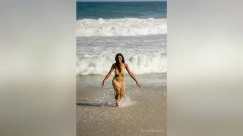 CrischelNovaks stands in the shallow ocean waves wearing a long, tan, open-front knit dress.