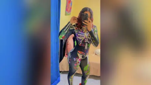 Young woman in a colorful psychedelic patterned bodysuit taking a mirror selfie in a bright bedroom.